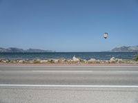 kites fly over the beach with a distant lake and mountains in the background from a highway