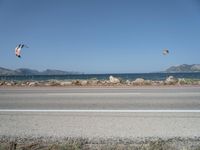 kites fly over the beach with a distant lake and mountains in the background from a highway