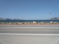 kites fly over the beach with a distant lake and mountains in the background from a highway