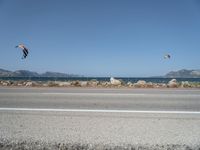 kites fly over the beach with a distant lake and mountains in the background from a highway