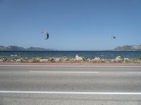 kites fly over the beach with a distant lake and mountains in the background from a highway