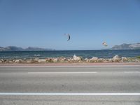 kites fly over the beach with a distant lake and mountains in the background from a highway