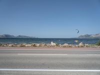 kites fly over the beach with a distant lake and mountains in the background from a highway