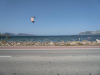 kites fly over the beach with a distant lake and mountains in the background from a highway