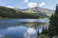 a large lake surrounded by some evergreen forest with a blue sky and white clouds above it