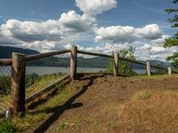 a fence that is next to some water near a field and trees on a hill