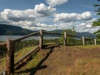 a fence that is next to some water near a field and trees on a hill