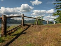 a fence that is next to some water near a field and trees on a hill