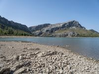 a body of water near some hills and trees with rocks on the shore of it