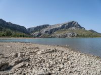 a body of water near some hills and trees with rocks on the shore of it