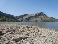 a body of water near some hills and trees with rocks on the shore of it