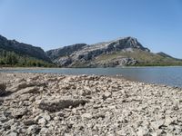 a body of water near some hills and trees with rocks on the shore of it