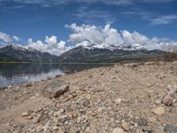 a lake with rocks on the shore and mountains in the background along side it and snow capped hills