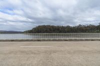 a fence in an empty parking lot overlooking water and trees of some type that is not native