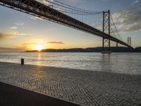 the golden hour lights up the sky near a long bridge over the water and a small area with squares on the ground in front