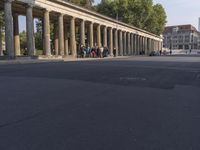 an empty street lined with pillars and a building in the back ground is a large group of people walking along it