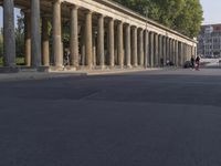 an empty street lined with pillars and a building in the back ground is a large group of people walking along it