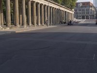 an empty street lined with pillars and a building in the back ground is a large group of people walking along it