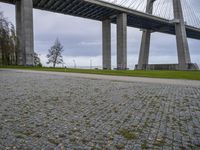 a large bridge spanning over a park with grass and brick sidewalking area below it