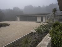 large garage with two buildings, trees and gravel area next to it, on a foggy day