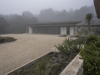 large garage with two buildings, trees and gravel area next to it, on a foggy day