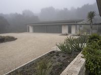 large garage with two buildings, trees and gravel area next to it, on a foggy day