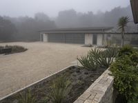 large garage with two buildings, trees and gravel area next to it, on a foggy day