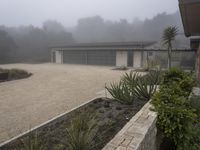 large garage with two buildings, trees and gravel area next to it, on a foggy day