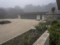 large garage with two buildings, trees and gravel area next to it, on a foggy day