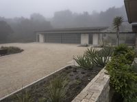 large garage with two buildings, trees and gravel area next to it, on a foggy day