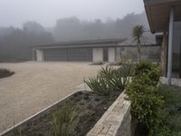 large garage with two buildings, trees and gravel area next to it, on a foggy day