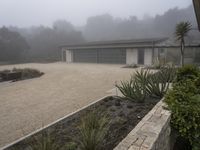 large garage with two buildings, trees and gravel area next to it, on a foggy day