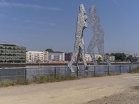 large metal sculptures of people facing each other on dirt path next to building complex with large metal structures in background