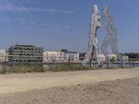 large metal sculptures of people facing each other on dirt path next to building complex with large metal structures in background