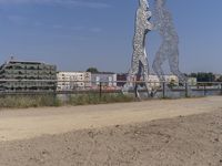 large metal sculptures of people facing each other on dirt path next to building complex with large metal structures in background