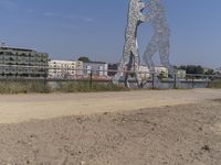 large metal sculptures of people facing each other on dirt path next to building complex with large metal structures in background