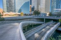 a curved walkway in front of a river in a city with tall buildings and tall trees