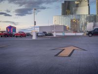 the parking lot has an arrow sign on it, near two buildings and a park