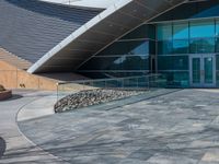 some concrete building with large glass windows and stone walkways outside of them with benches on the sidewalk next to the doors