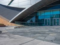 some concrete building with large glass windows and stone walkways outside of them with benches on the sidewalk next to the doors