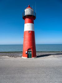 a tall red and white lighthouse on a beach next to the ocean with a blue sky