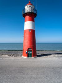 a tall red and white lighthouse on a beach next to the ocean with a blue sky