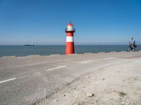 an orange and white lighthouse is next to the ocean near some cars and people on the road