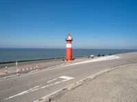 a lighthouse sits on a road next to the ocean and a beach with cars and bikes