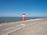 a lighthouse sits on a road next to the ocean and a beach with cars and bikes