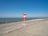 a lighthouse sits on a road next to the ocean and a beach with cars and bikes