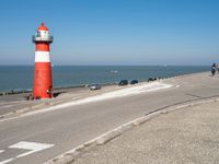 a lighthouse sits on a road next to the ocean and a beach with cars and bikes