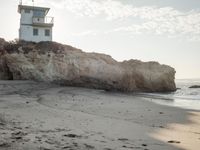 a lighthouse stands high on a rocky cliff at the beach with footprints in the sand