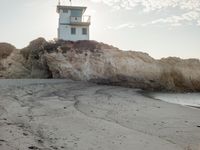 a lighthouse stands high on a rocky cliff at the beach with footprints in the sand