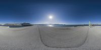 a fisheye lens photo of a beach with snow capped mountains in the distance and people on a bench, with sun reflecting off